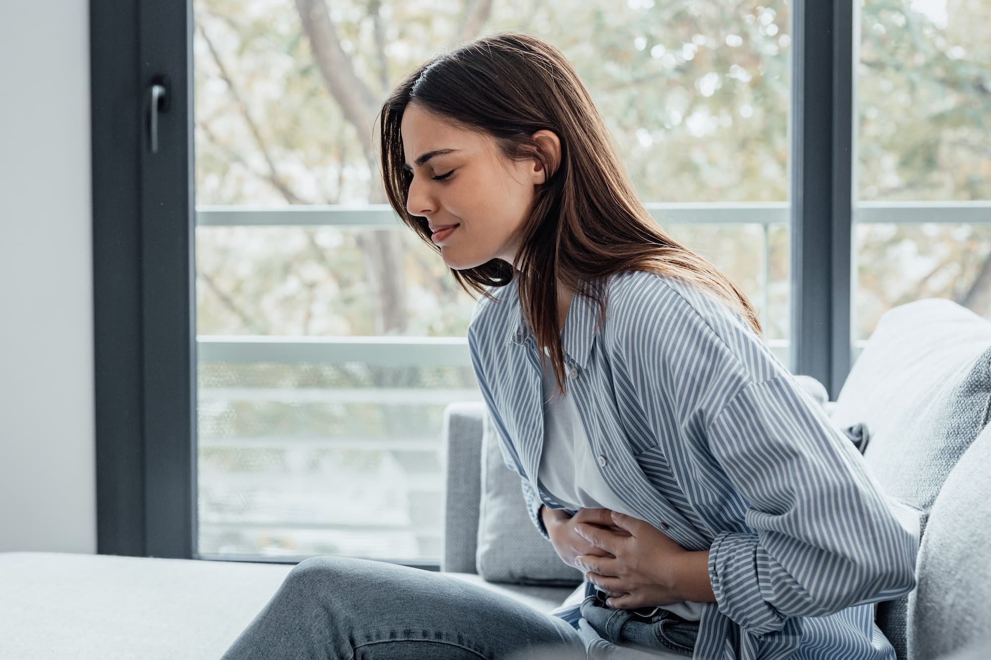 Young woman sitting on a couch and clutching her stomach to cope with her pelvic discomfort near Plano, TX.