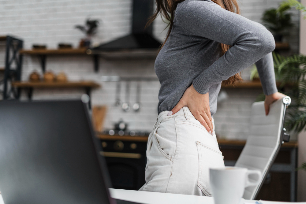 A woman standing near her desk with her hand on her lower back in a kitchen as she considers calling a lower back specialist near Plano.